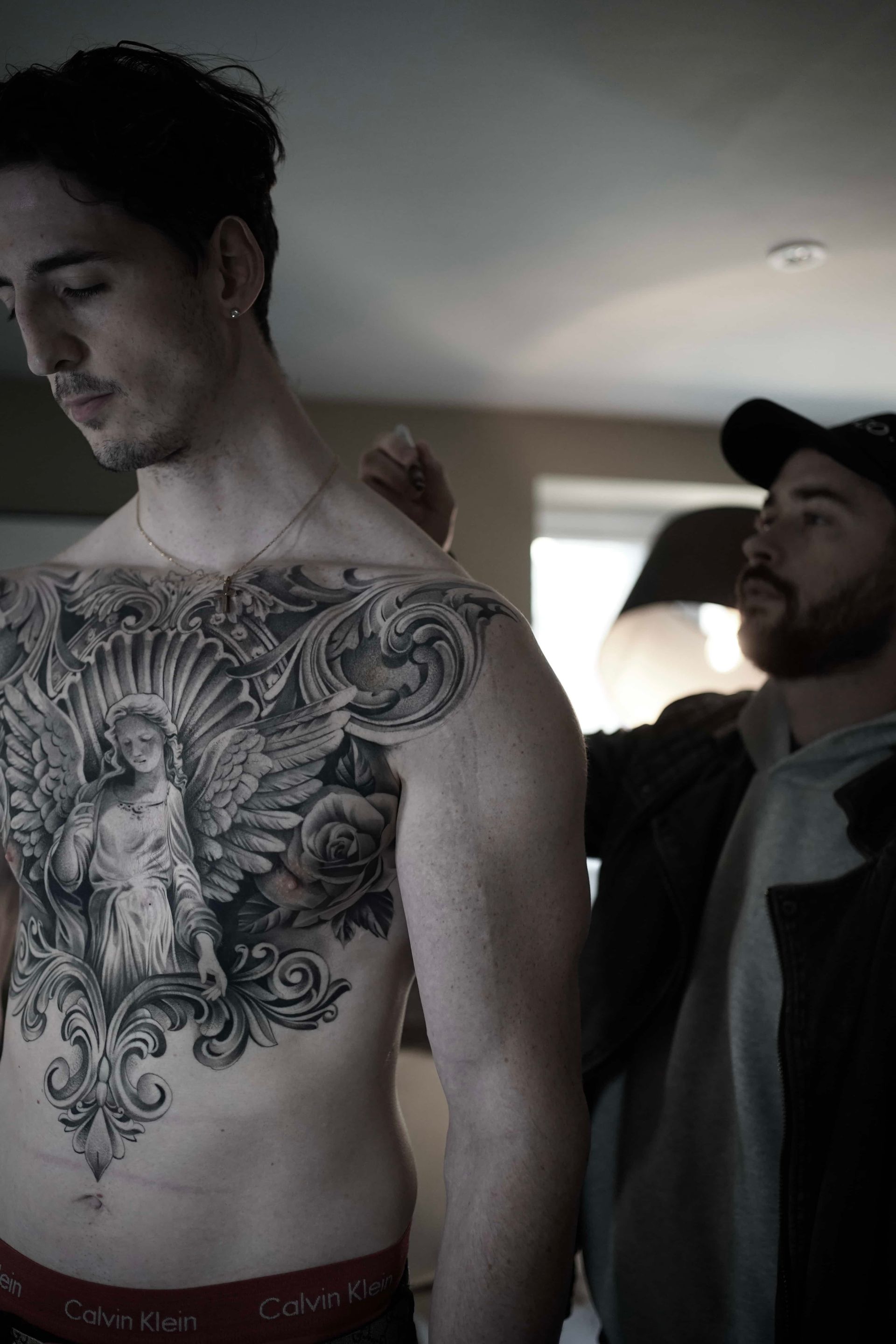 Man with large chest tattoo being prepped by another person, indoors.