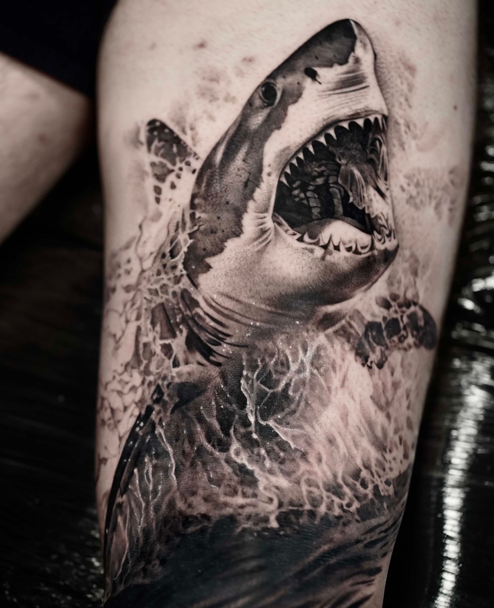 Tattoo of a great white shark breaching the water, mouth open, with grayscale details.