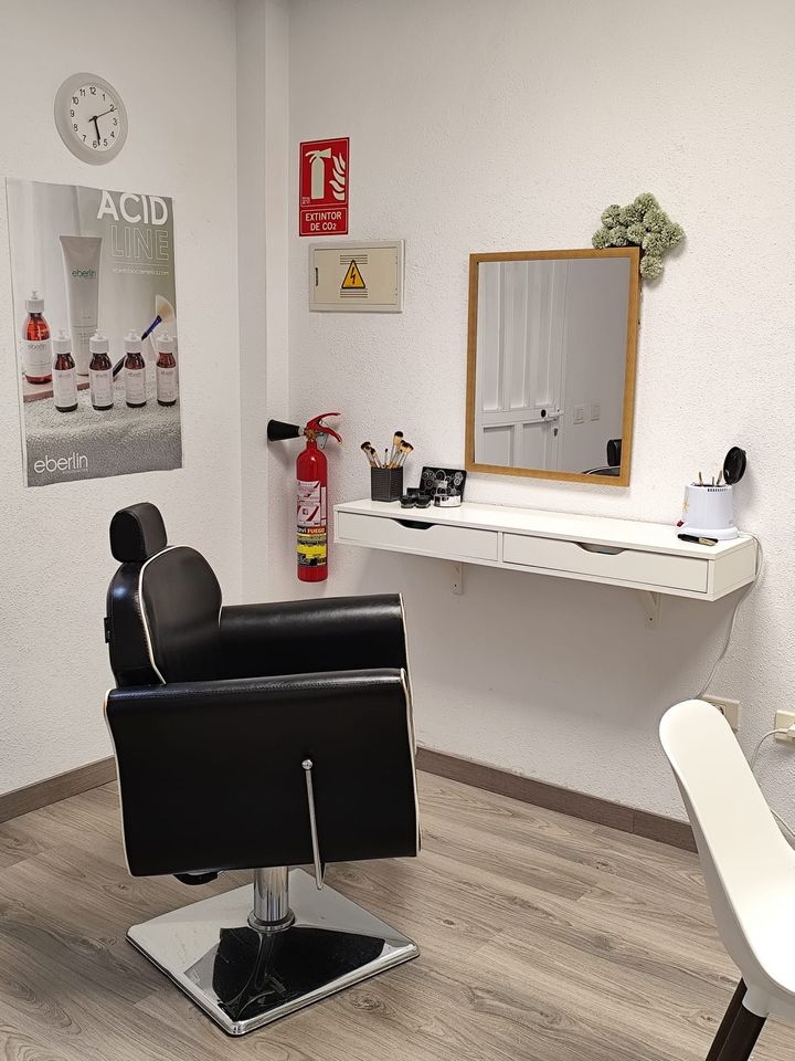 Rincón de peluquería: silla de peluquería negra, estante flotante blanco con espejo, cosméticos y un extintor de incendios en una pared texturizada.