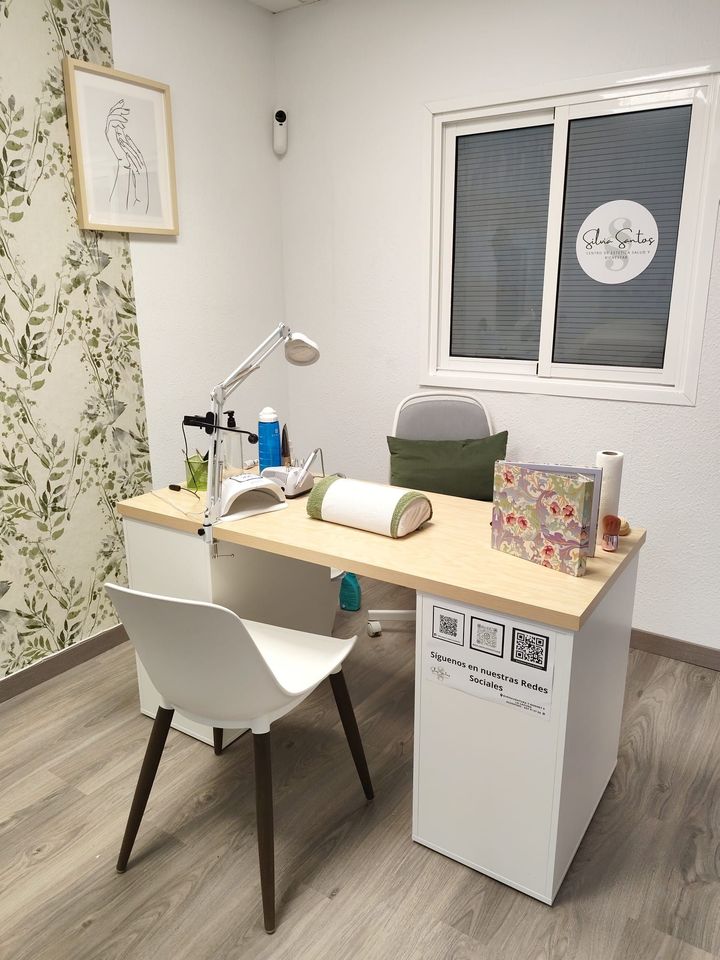 Una estación de manicura en una habitación bien iluminada, con un escritorio, una silla blanca y herramientas. La pared de la izquierda tiene papel pintado floral.