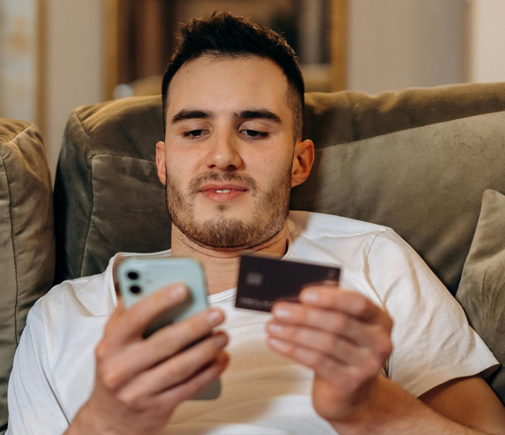 A man is sitting on a couch holding a credit card and a cell phone.