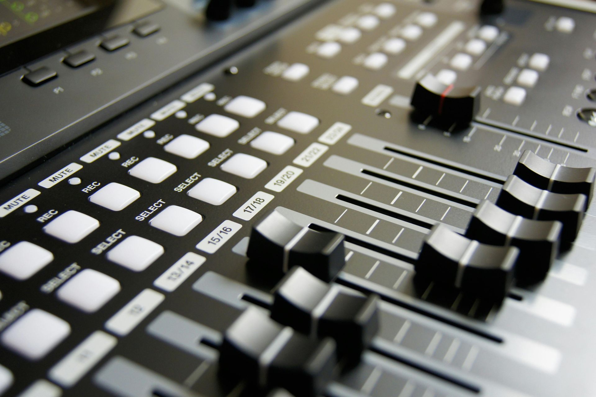 Close-up of a mixing console with sliders and buttons, angled view, grayscale color scheme.