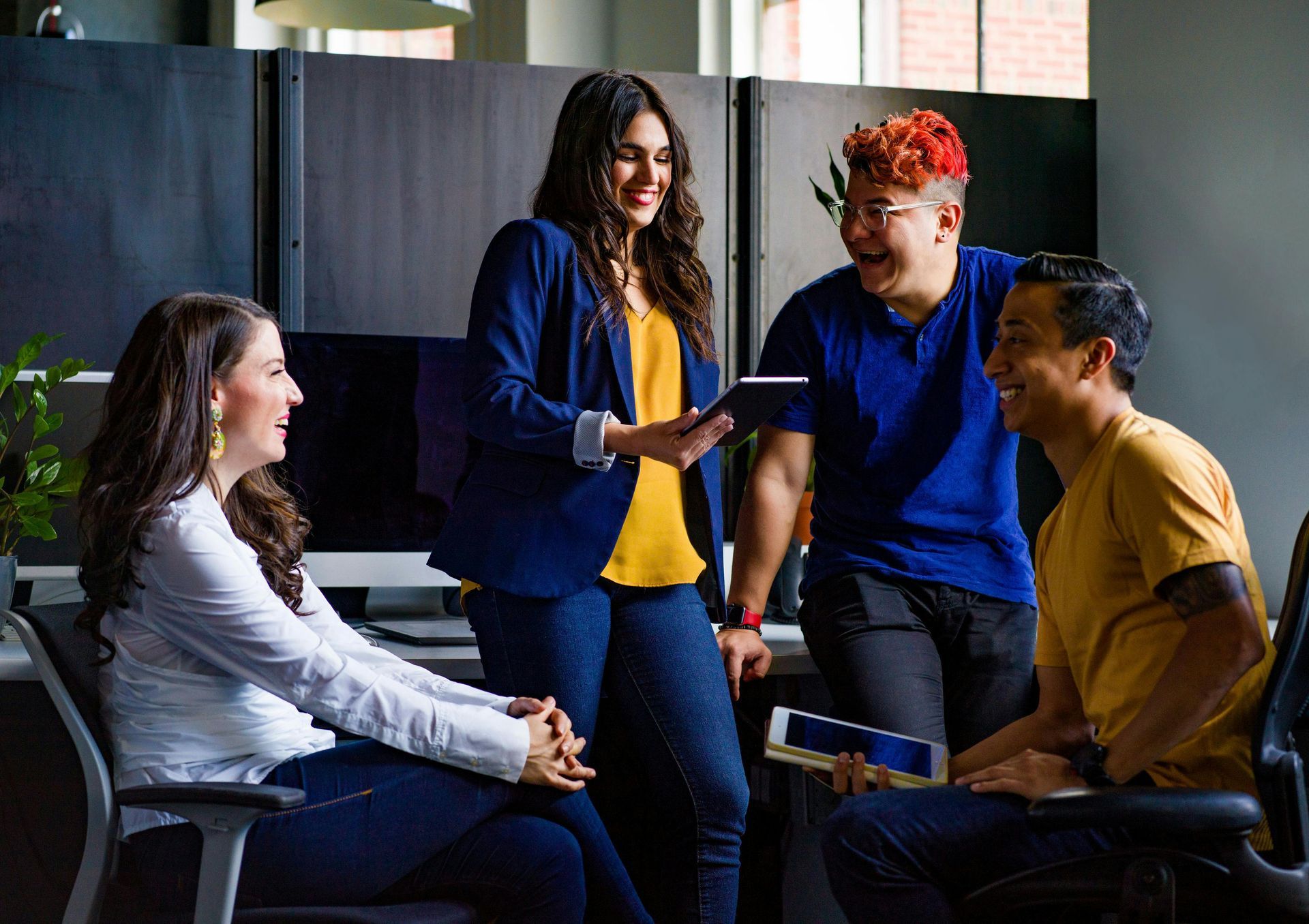 Four people in an office setting laugh and talk while looking at tablets.