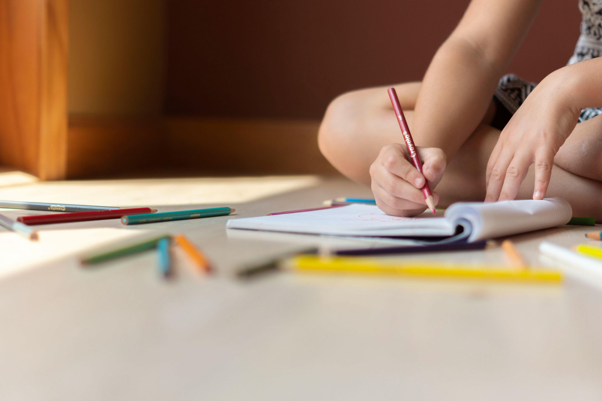 Child sitting on floor, drawing with pencil in a notebook, surrounded by colored pencils.