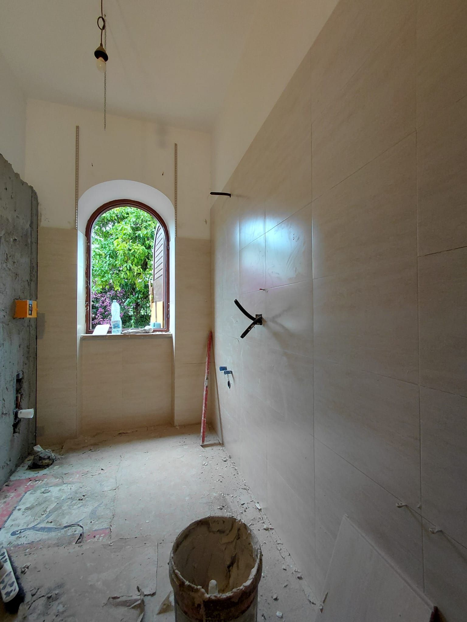 Ristrutturazione in corso in un bagno stretto con pareti piastrellate, finestra ad arco e secchio da cantiere sul pavimento.