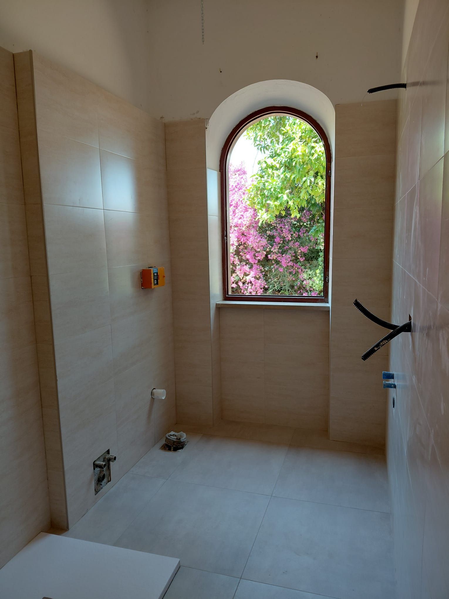Bagno con doccia piastrellato e finestra ad arco che si affaccia su fiori rosa e vegetazione verde.