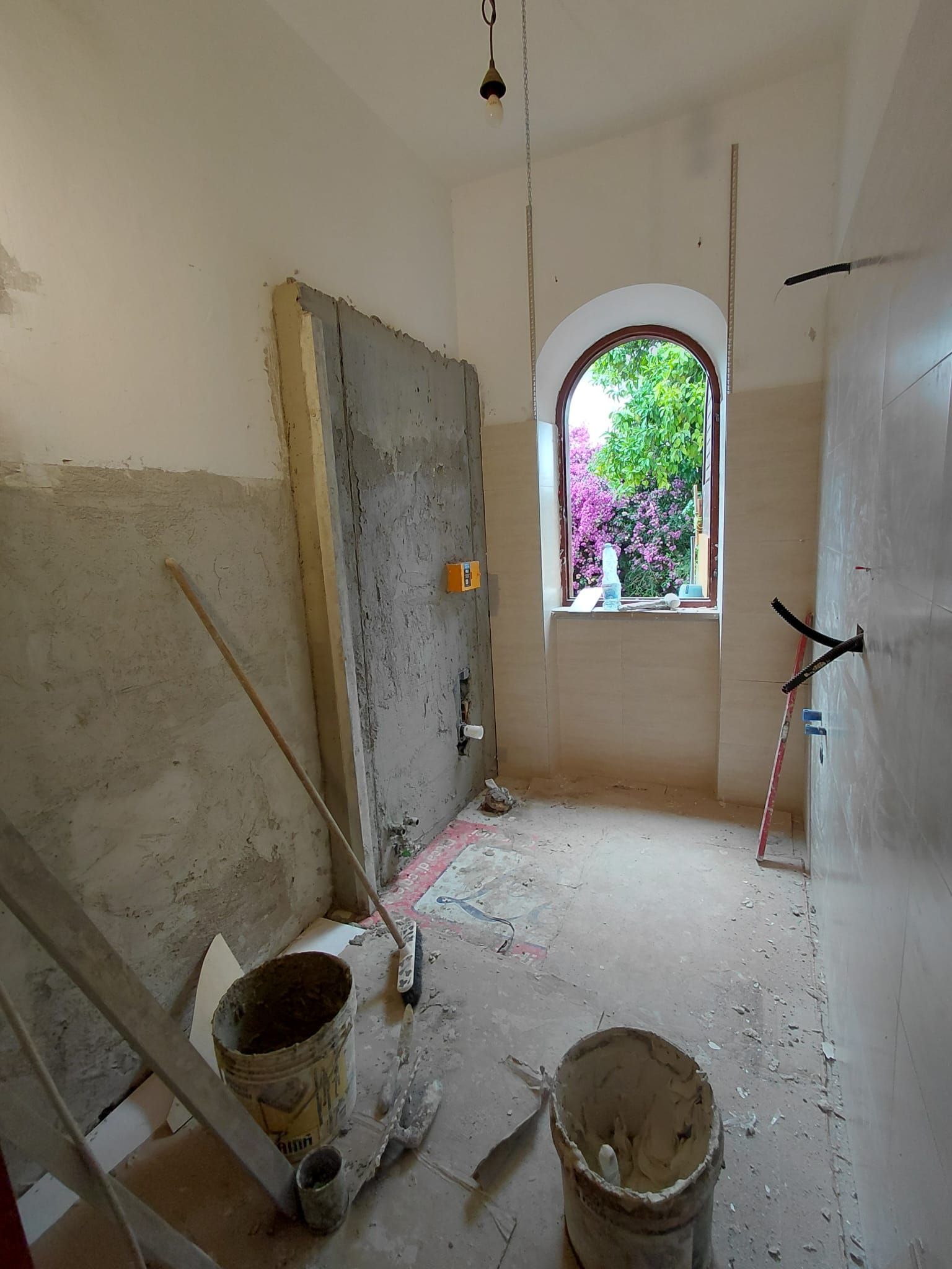 Stanza in ristrutturazione con pareti intonacate, secchi e una finestra che si affaccia sui fiori.