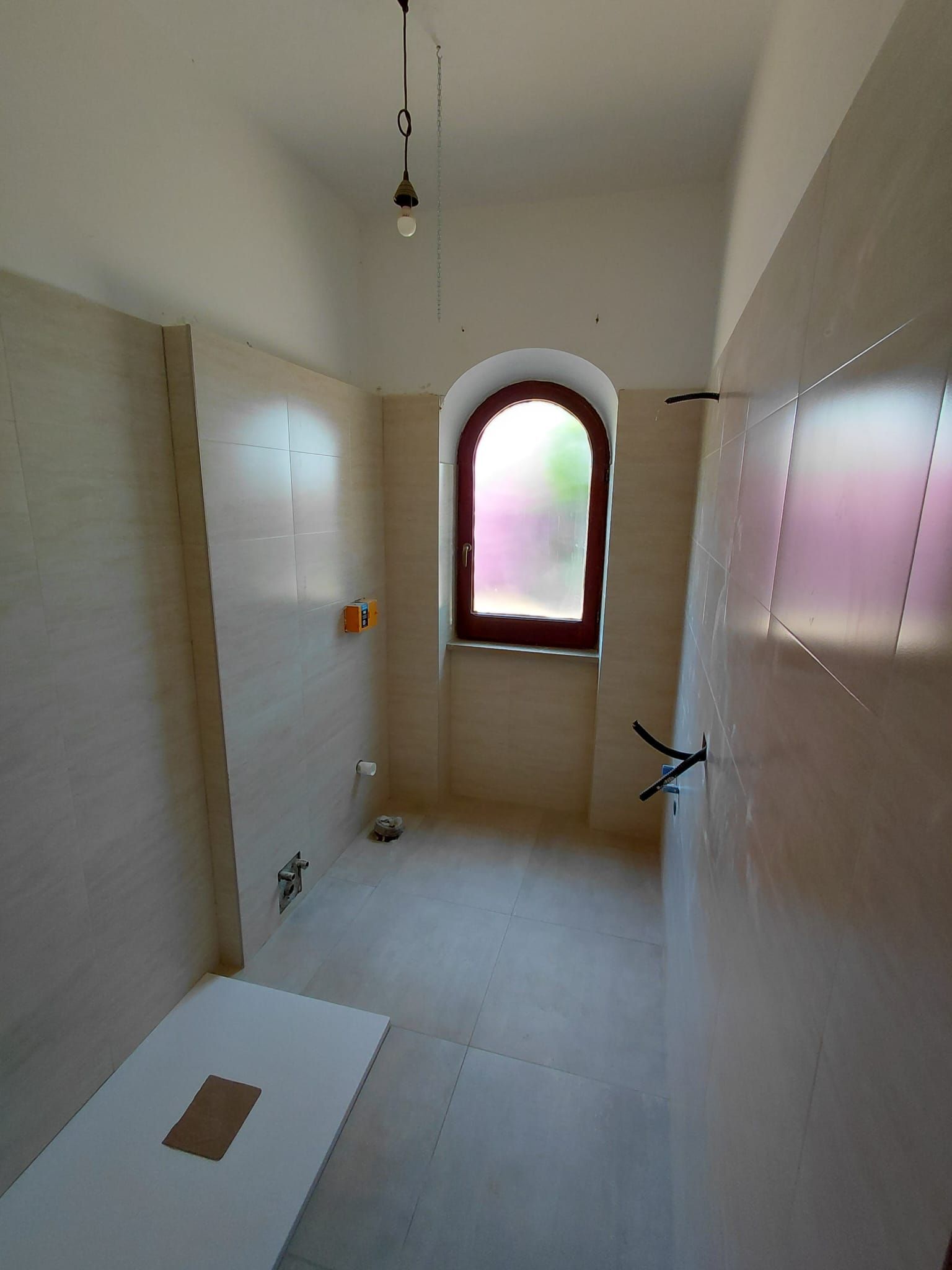 Bagno stretto piastrellato con finestra ad arco satinata, rubinetteria per la doccia e base del WC visibili in primo piano.