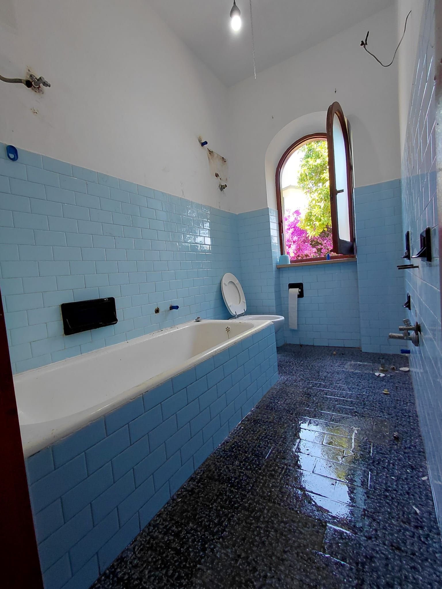 Bagno con piastrelle blu brillante, vasca bianca, pavimento scuro lucido e finestra ad arco con vetri colorati.