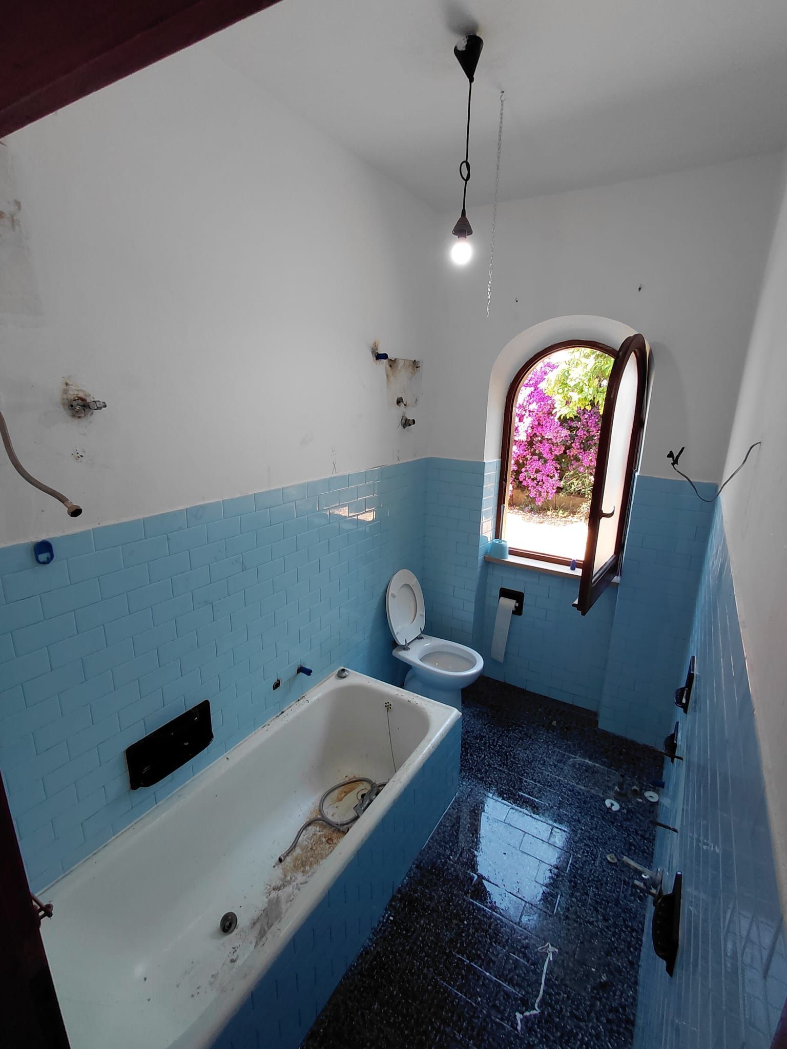 Piccolo bagno con pareti rivestite di piastrelle blu, vasca da bagno, WC e una finestra ad arco con fiori all'esterno.