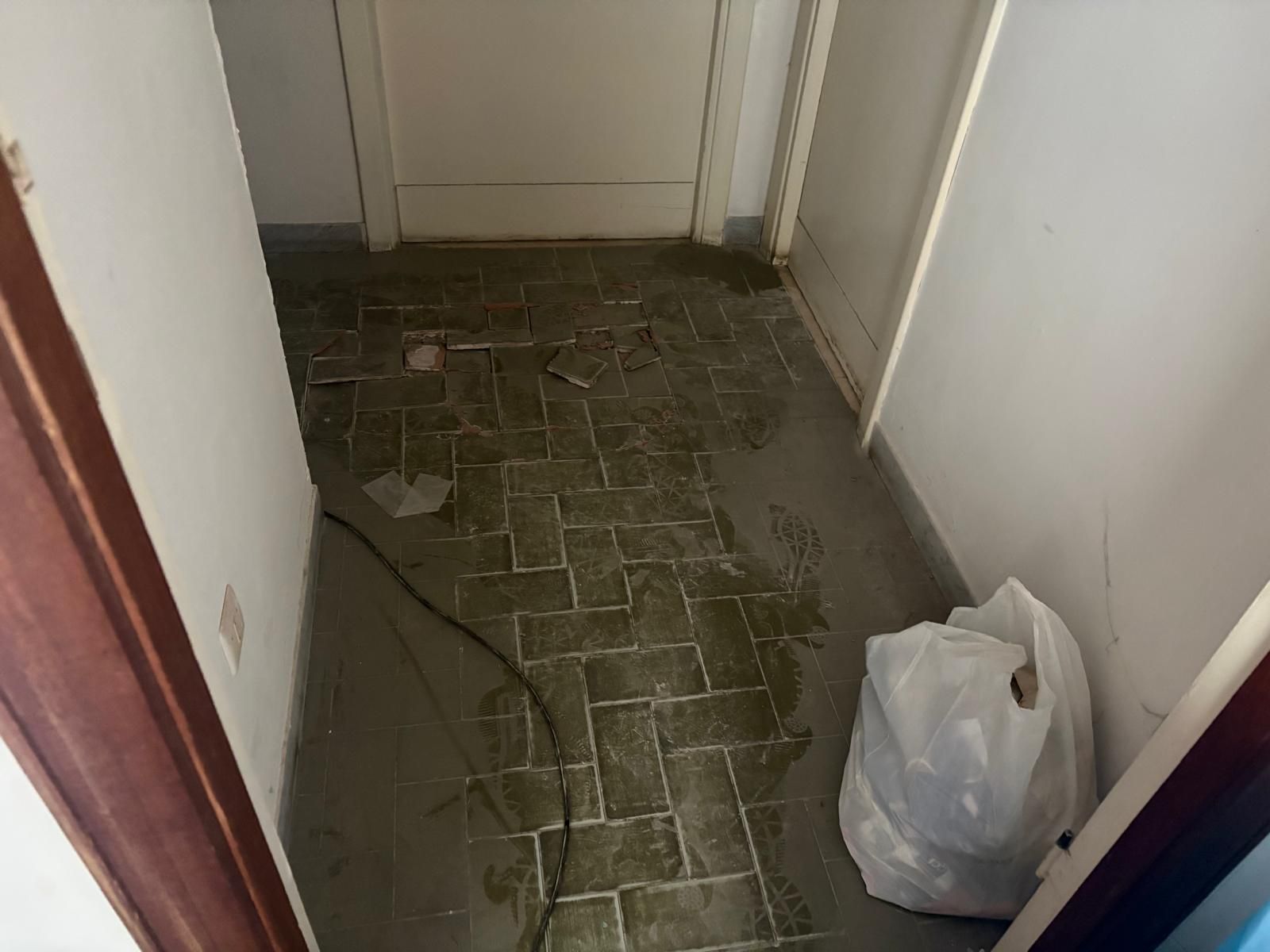 Stretto corridoio con pavimento piastrellato bagnato e sporco e un sacchetto di plastica bianco appoggiato al muro.