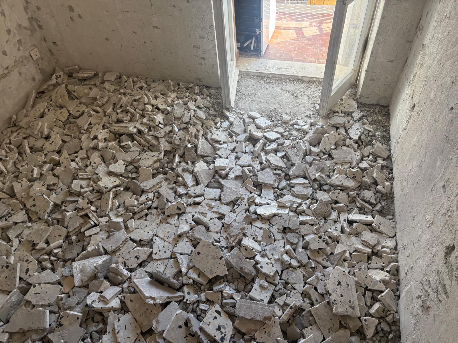 Stanza interna con pavimento ricoperto di frammenti di piastrelle bianche rotte, vicino a una porta aperta.