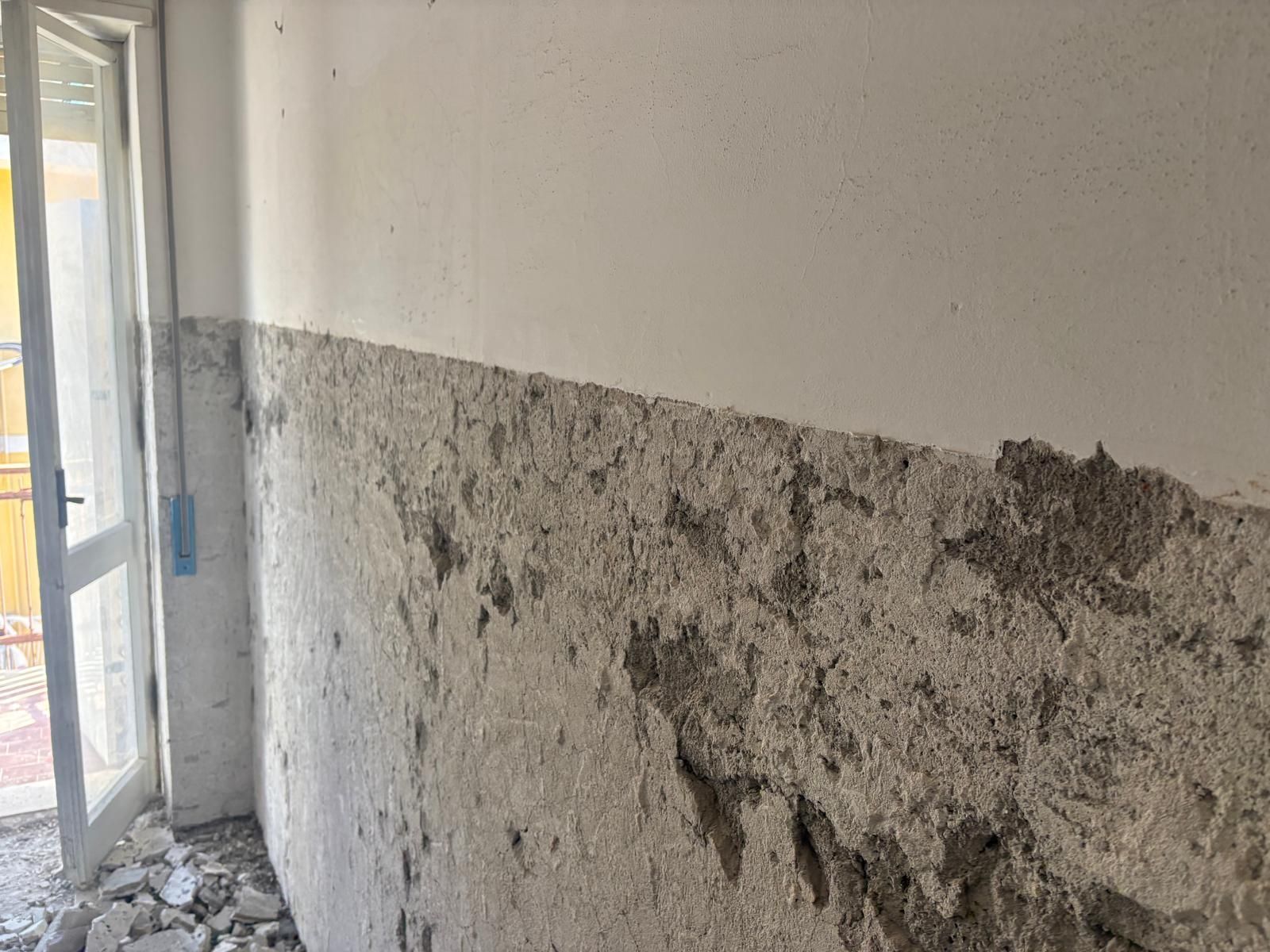 In un corridoio stretto, l'intonaco si scrosta, lasciando intravedere delle chiazze grigie e aprendo una porta sulla sinistra.