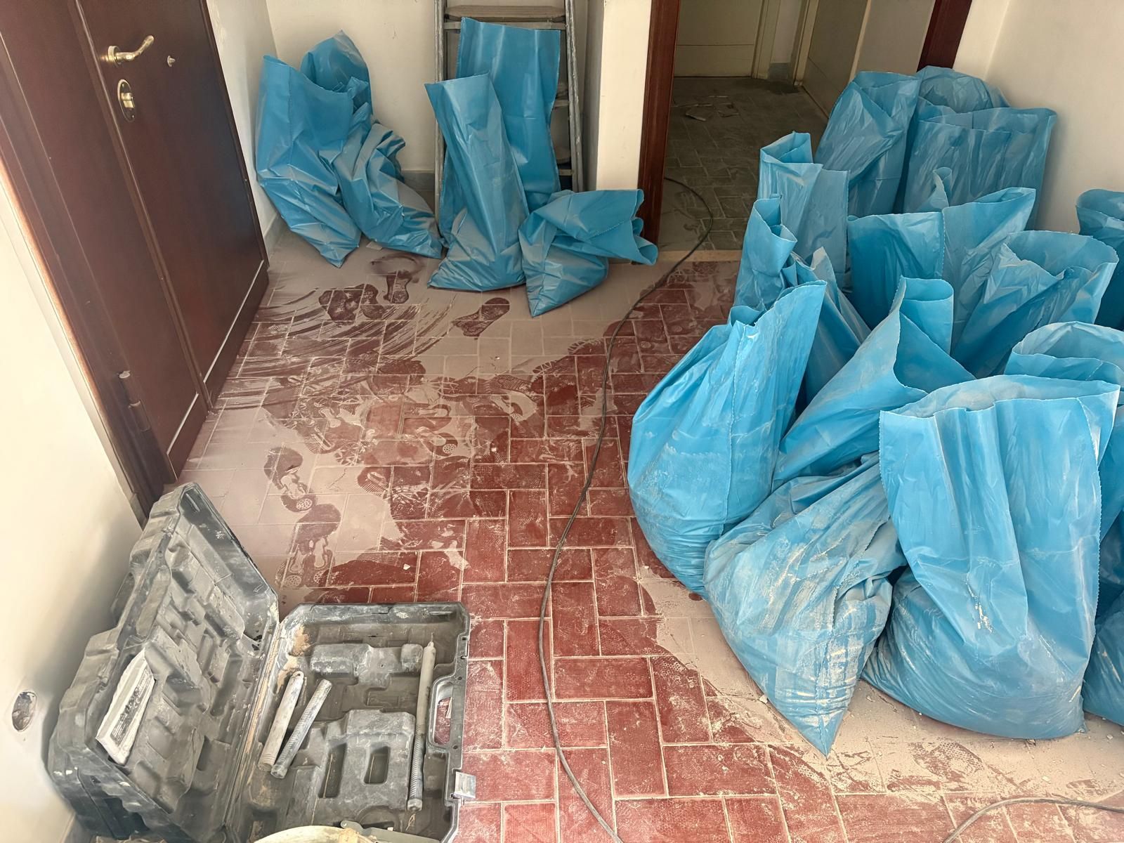 Borse blu e vassoi grigi impilati su un pavimento piastrellato in una piccola stanza o corridoio.