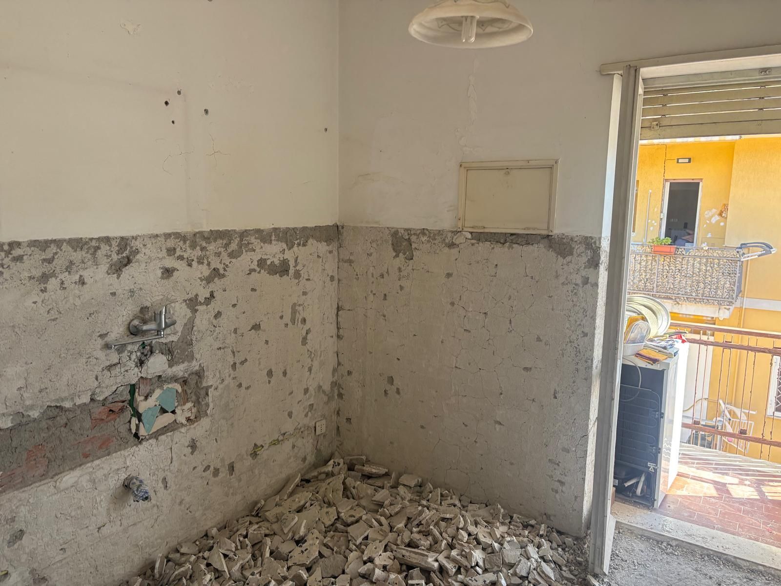 Cucina danneggiata con pavimento coperto di detriti, tubature a vista e una finestra che si apre su un esterno luminoso