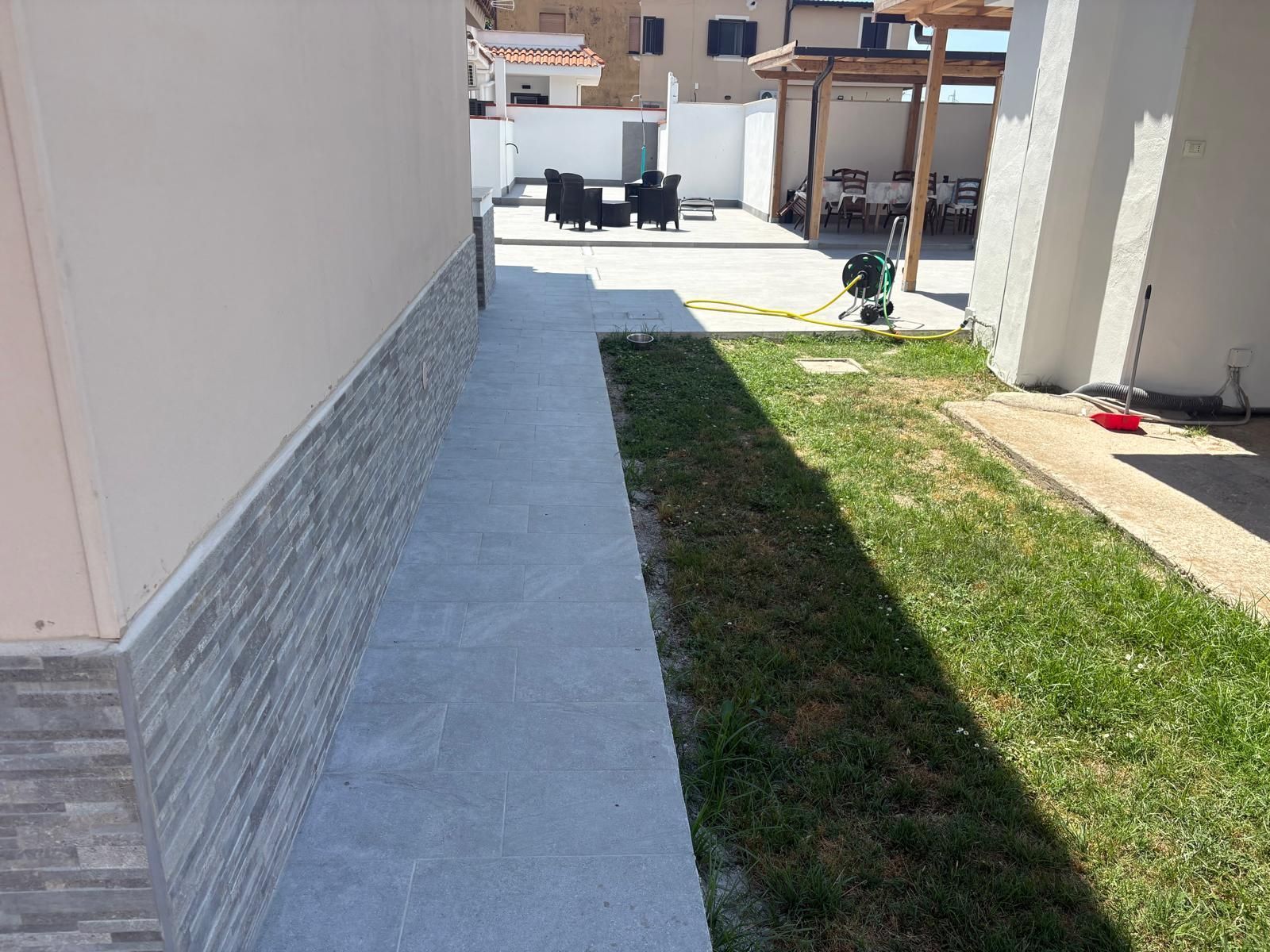 Stretto passaggio pedonale in cemento accanto a un edificio, con erba, un'ombra e un'area patio in lontananza.