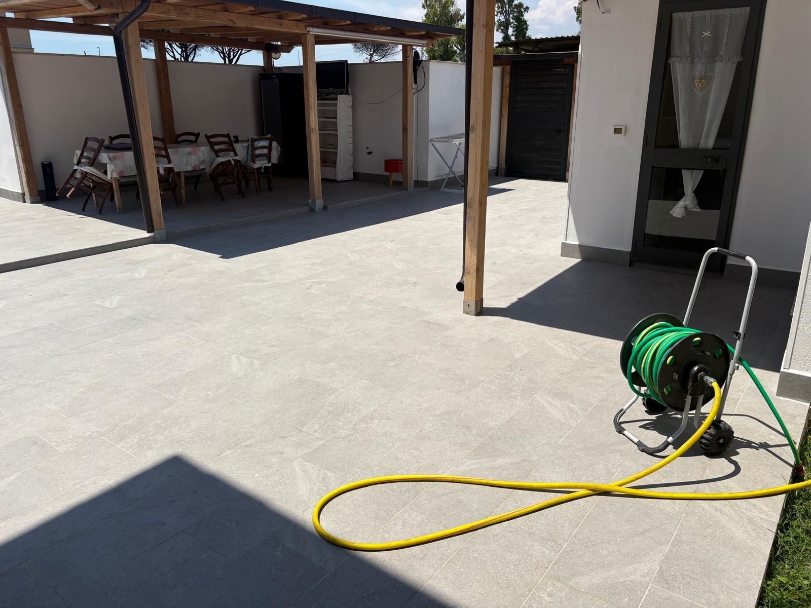 Patio con avvolgitubo verde e tubo giallo su una superficie di cemento illuminata dal sole.