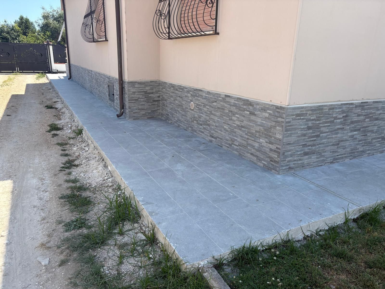 Passerella in cemento lungo il lato di una casa beige con base in pietra, accanto a un cortile sterrato e a chiazze d'erba.