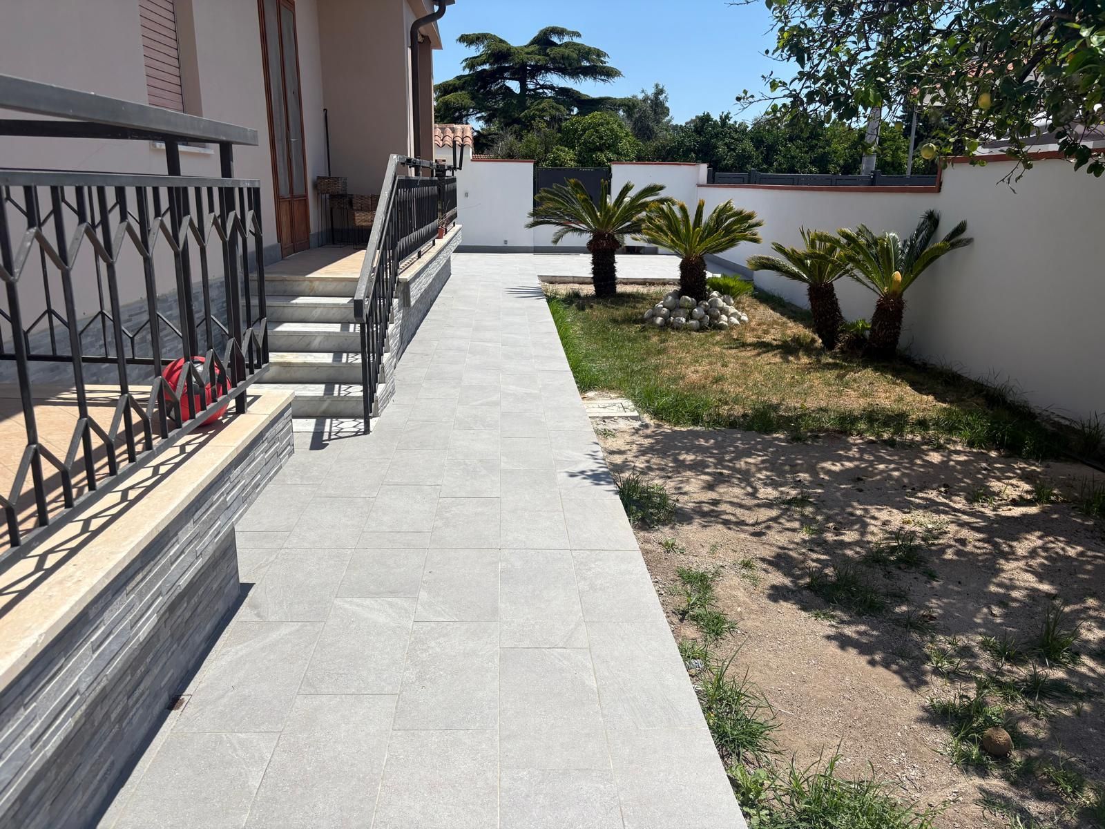 Passerella in cemento accanto a una casa con gradini, ringhiera e piccole piante di palma lungo un muro bianco