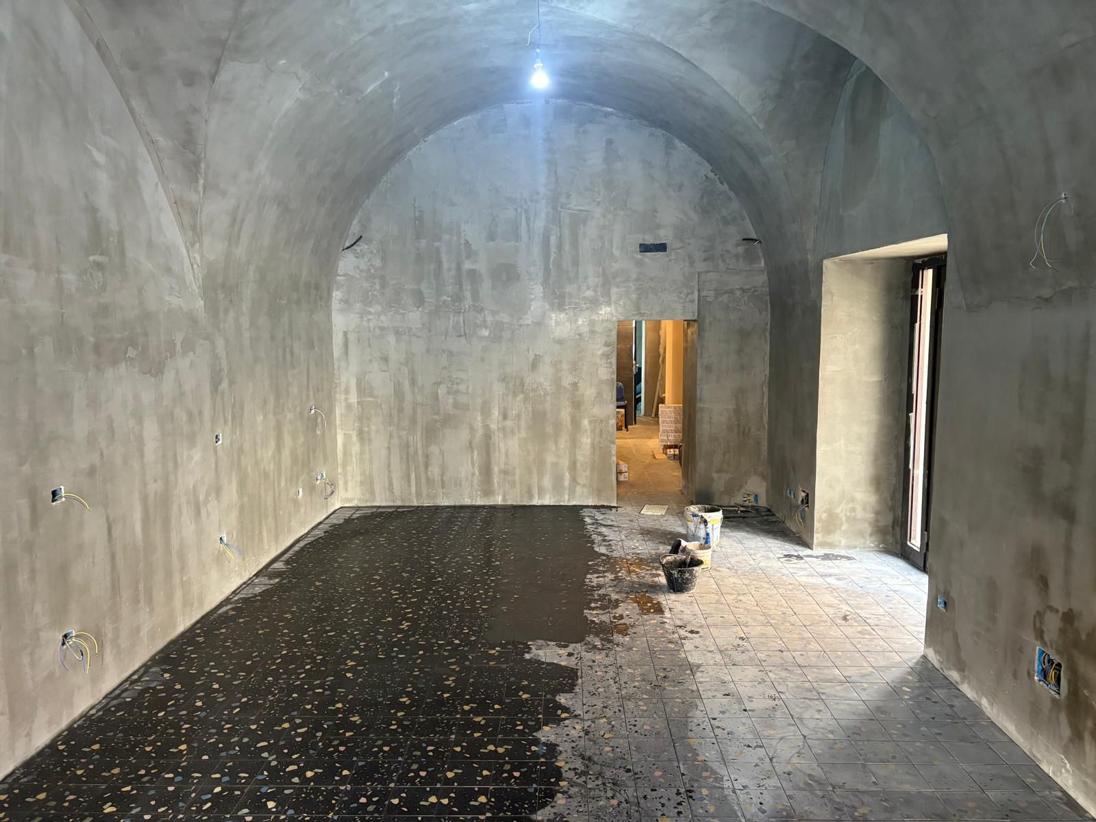 Interno in cemento a vista con soffitto a volta, pavimento bagnato e una porta aperta in fondo.
