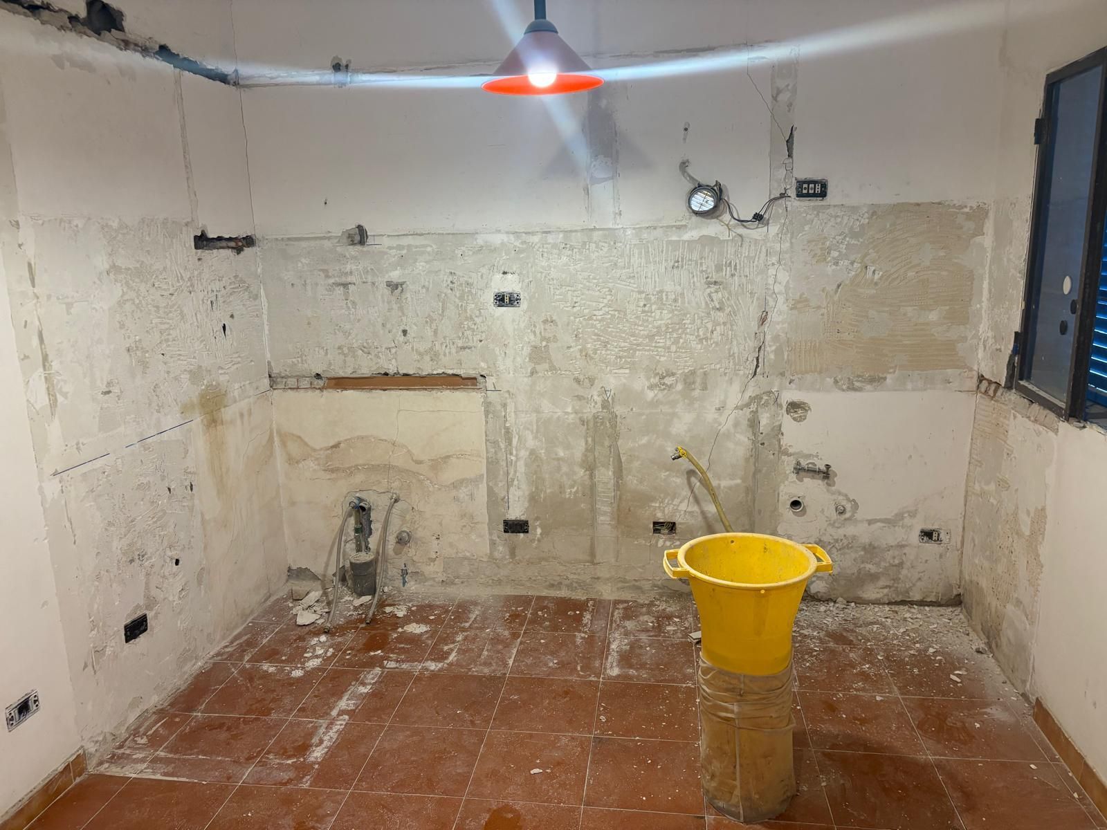 Stanza vuota e non rifinita con pavimento piastrellato, pareti scrostate, infissi a vista e un secchio giallo al centro.