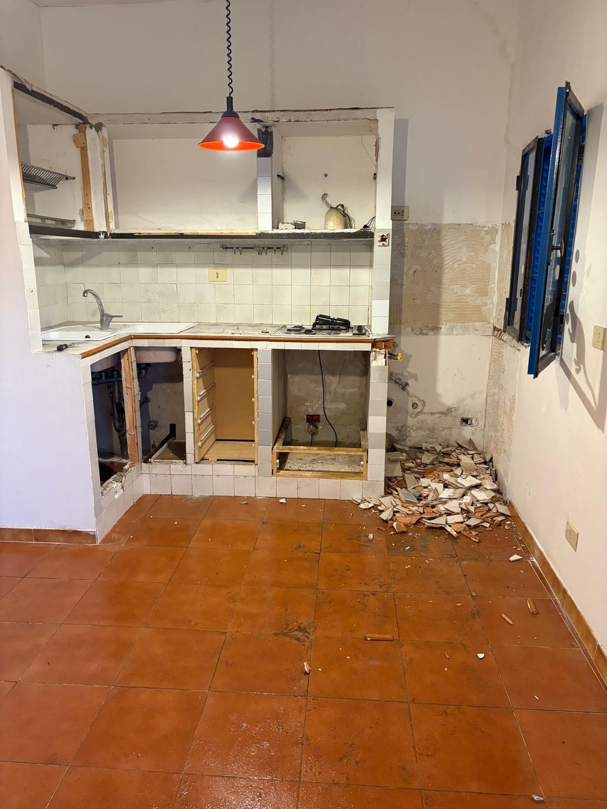 Cucina danneggiata con pensili mancanti, pavimento in piastrelle arancioni, lampada a sospensione mancante e detriti vicino alla finestra.