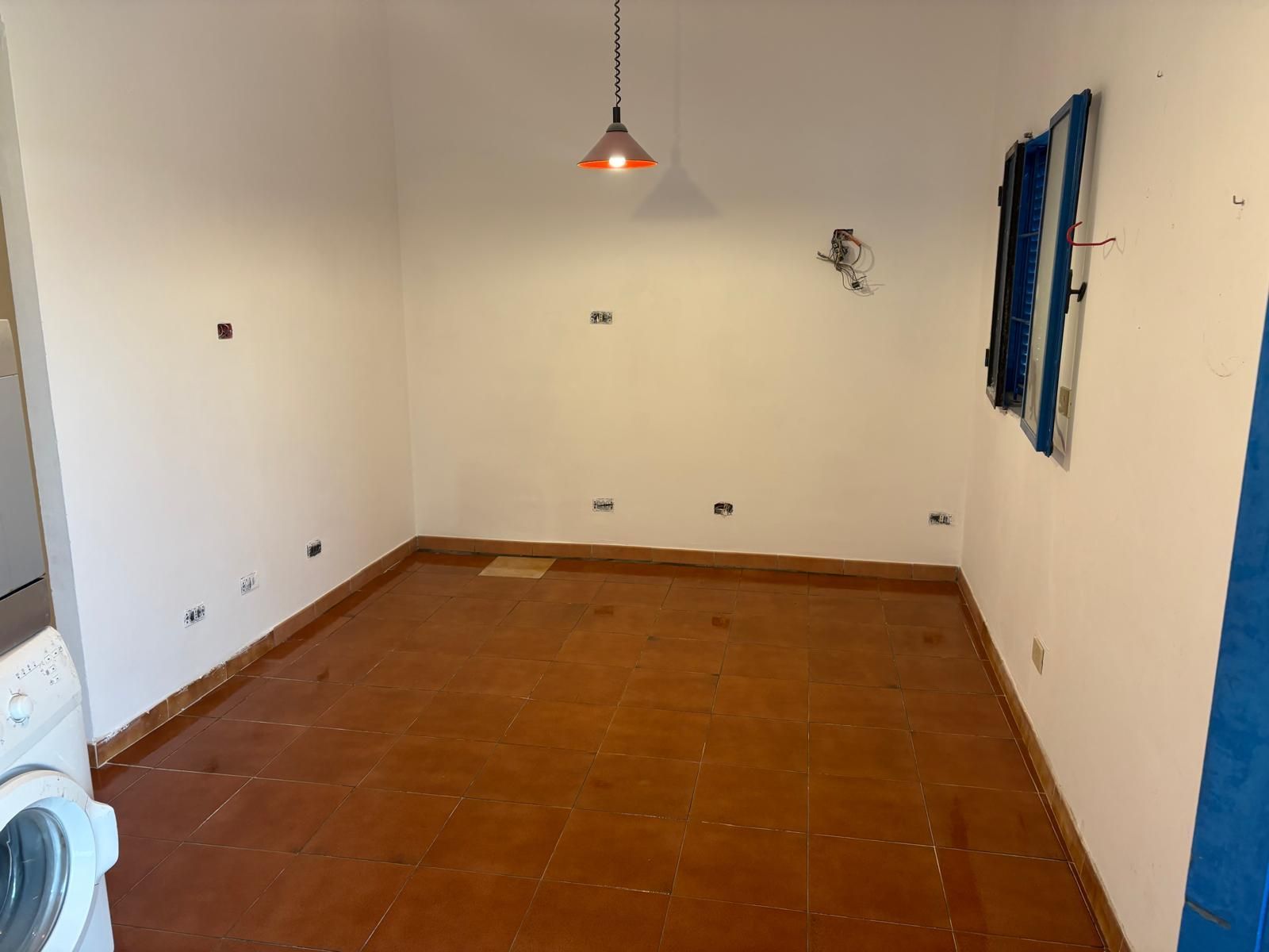 Stanza vuota con pavimento in piastrelle marroni, pareti bianche e lampadario a sospensione.