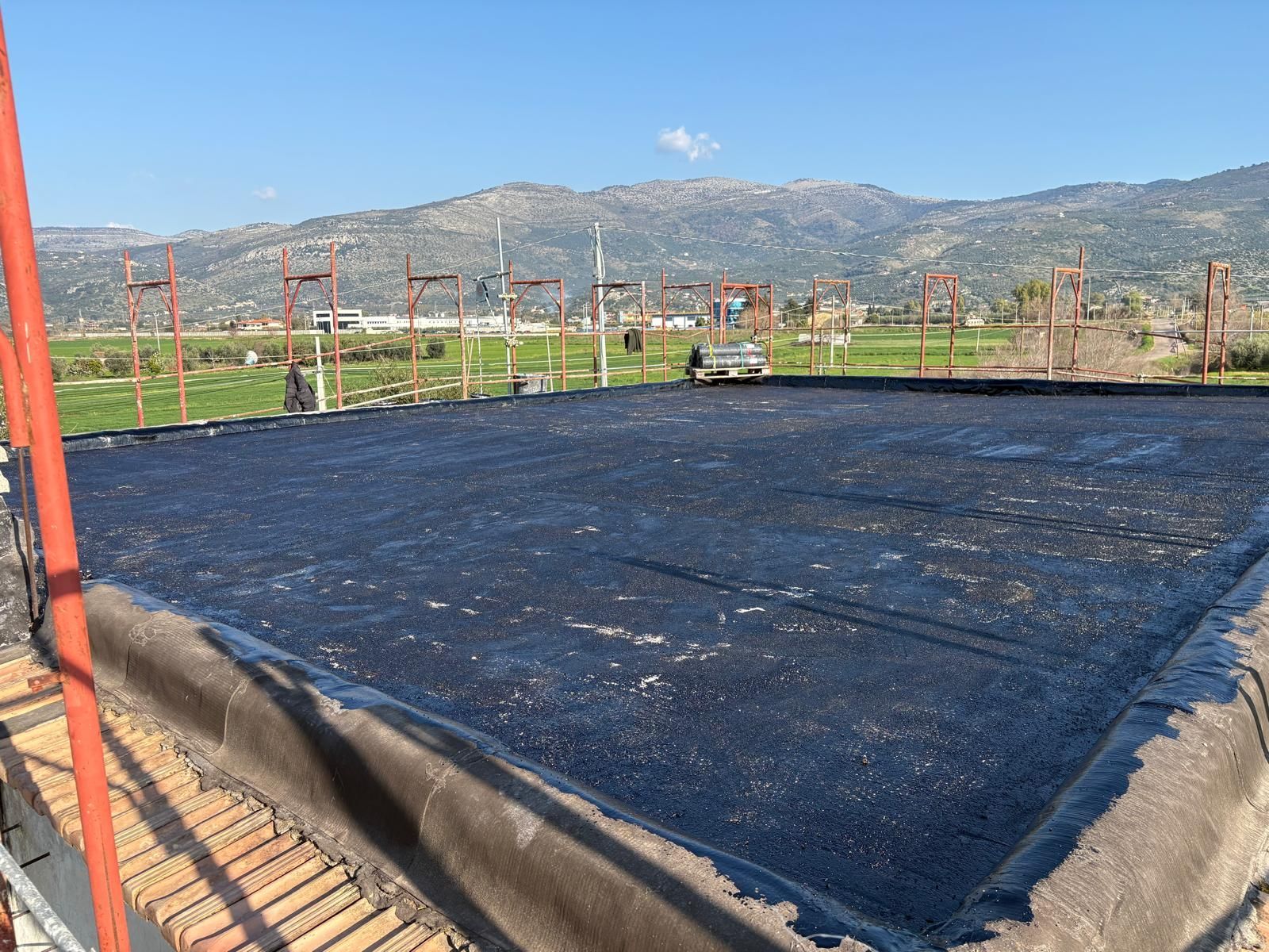 Un terreno scuro ricoperto da un telone, con pali di recinzione e montagne sotto un cielo azzurro limpido.