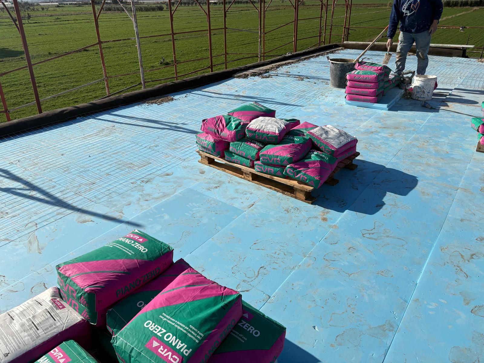 Pallet di materiali insacchettati su un telone blu ai margini di un campo, con un operaio nelle vicinanze.