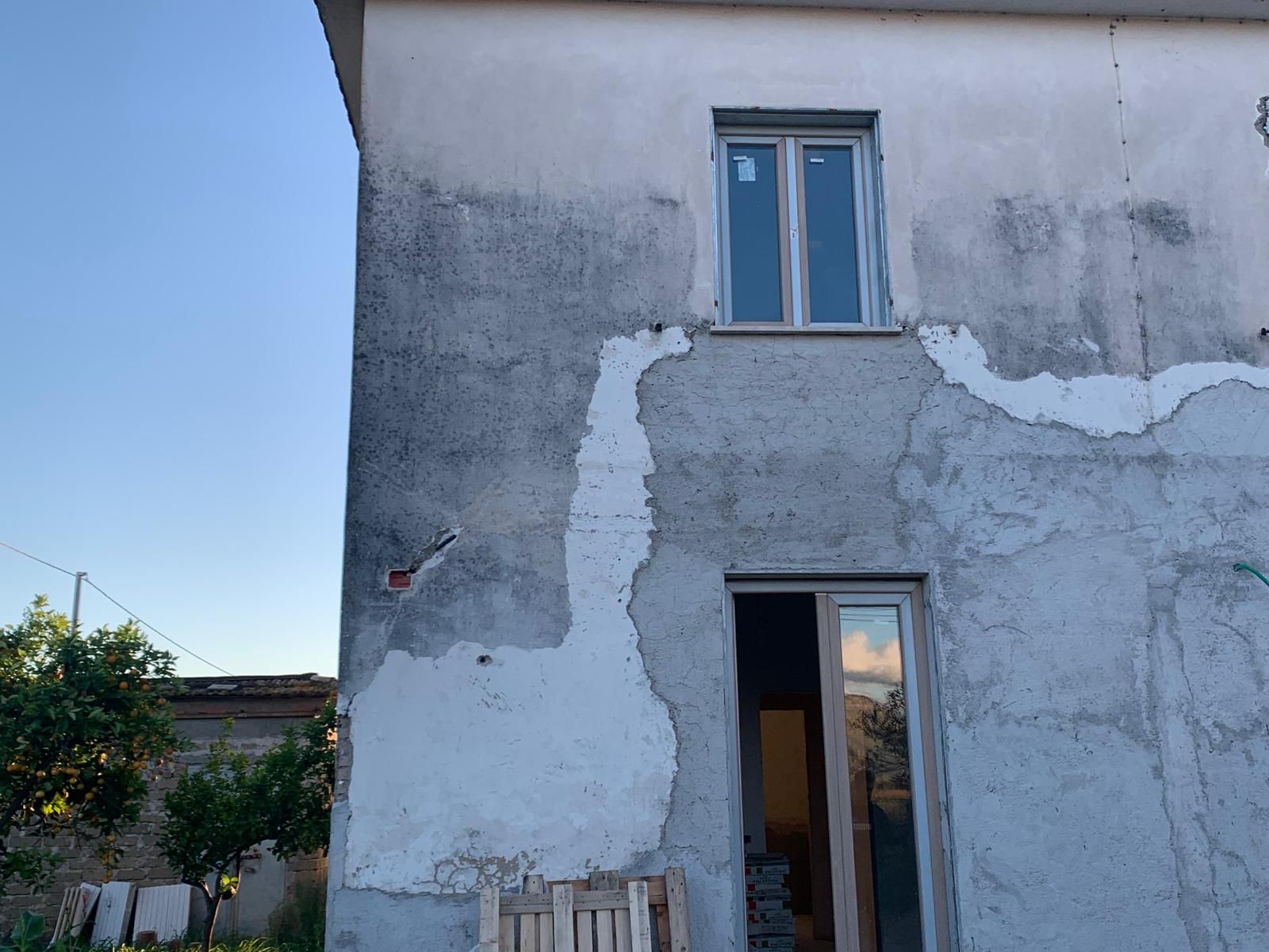 Esterno di una casa bianca e deteriorata dagli agenti atmosferici, con intonaco scrostato, finestra superiore blu e porta a vetri sottostante.