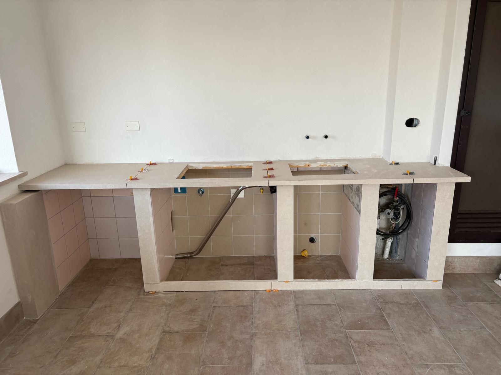 Cucina non rifinita con piano di lavoro bianco, ante a vista e tubature esposte.