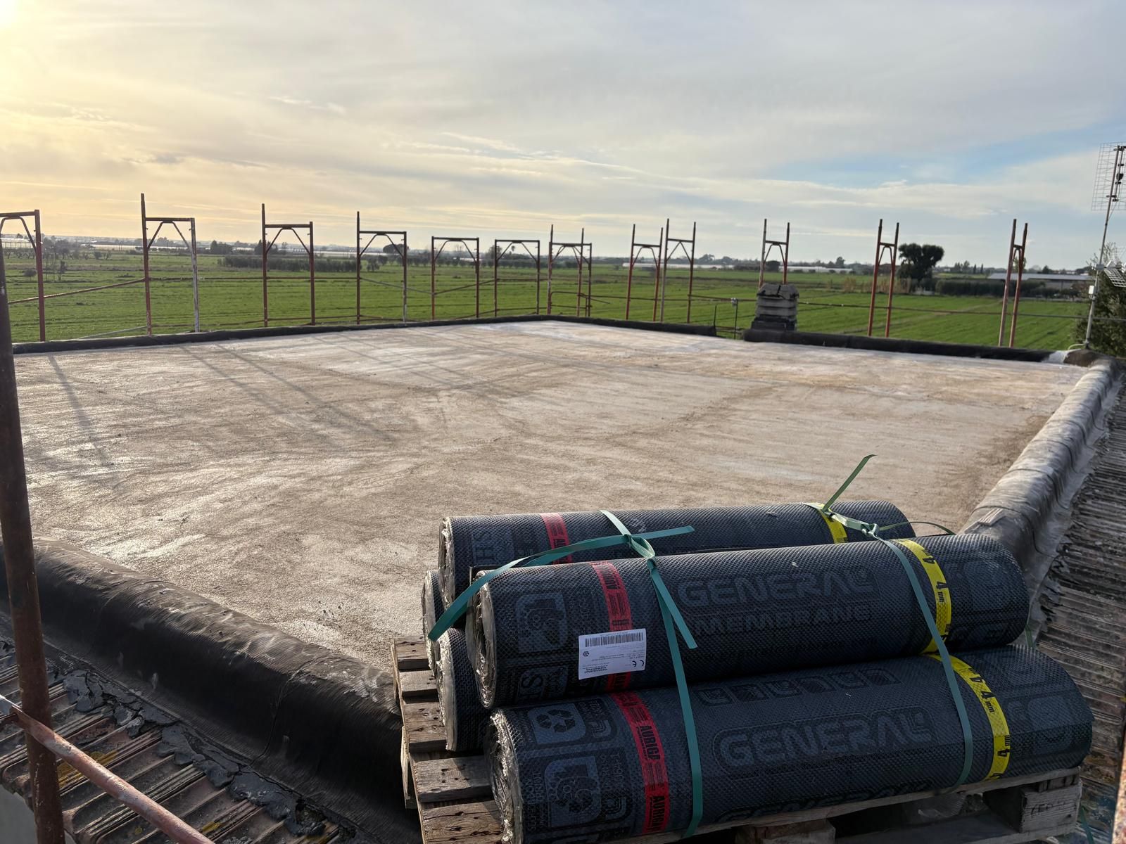In primo piano, in un cantiere recintato, si vede una nuova lastra di cemento con materiale di copertura in rotoli accatastato.