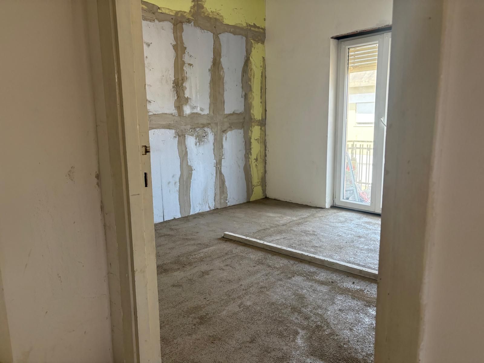 Stanza vuota ristrutturata con pareti non rifinite, pavimento in cemento e porta a vetri che dà su un balcone.