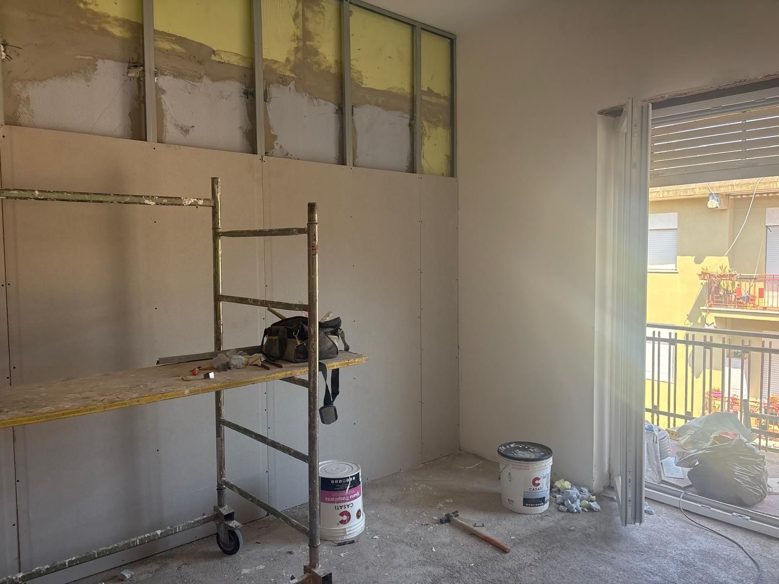 Interno di una stanza in fase di ristrutturazione con impalcature, attrezzi e struttura del muro a vista vicino a una finestra e a una porta-finestra.