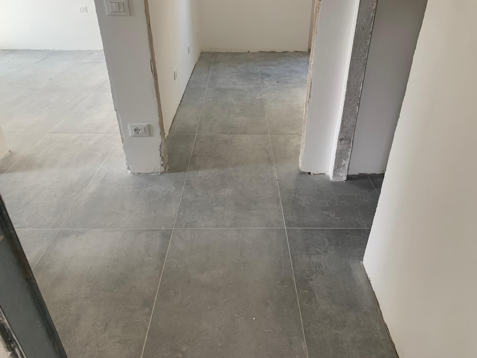 Corridoio interno vuoto con pavimento piastrellato grigio e pareti bianche, che lascia intravedere porte e cornici non rifinite.