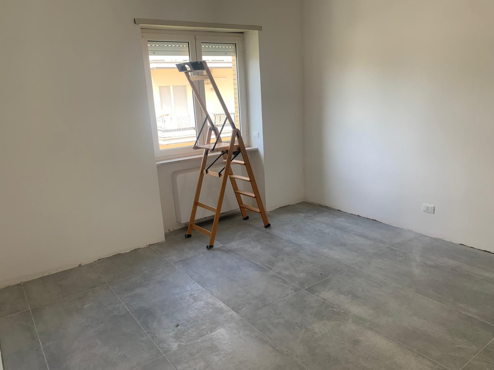 Stanza vuota con pareti e pavimento spogli, scala accanto a una finestra in fase di ristrutturazione.