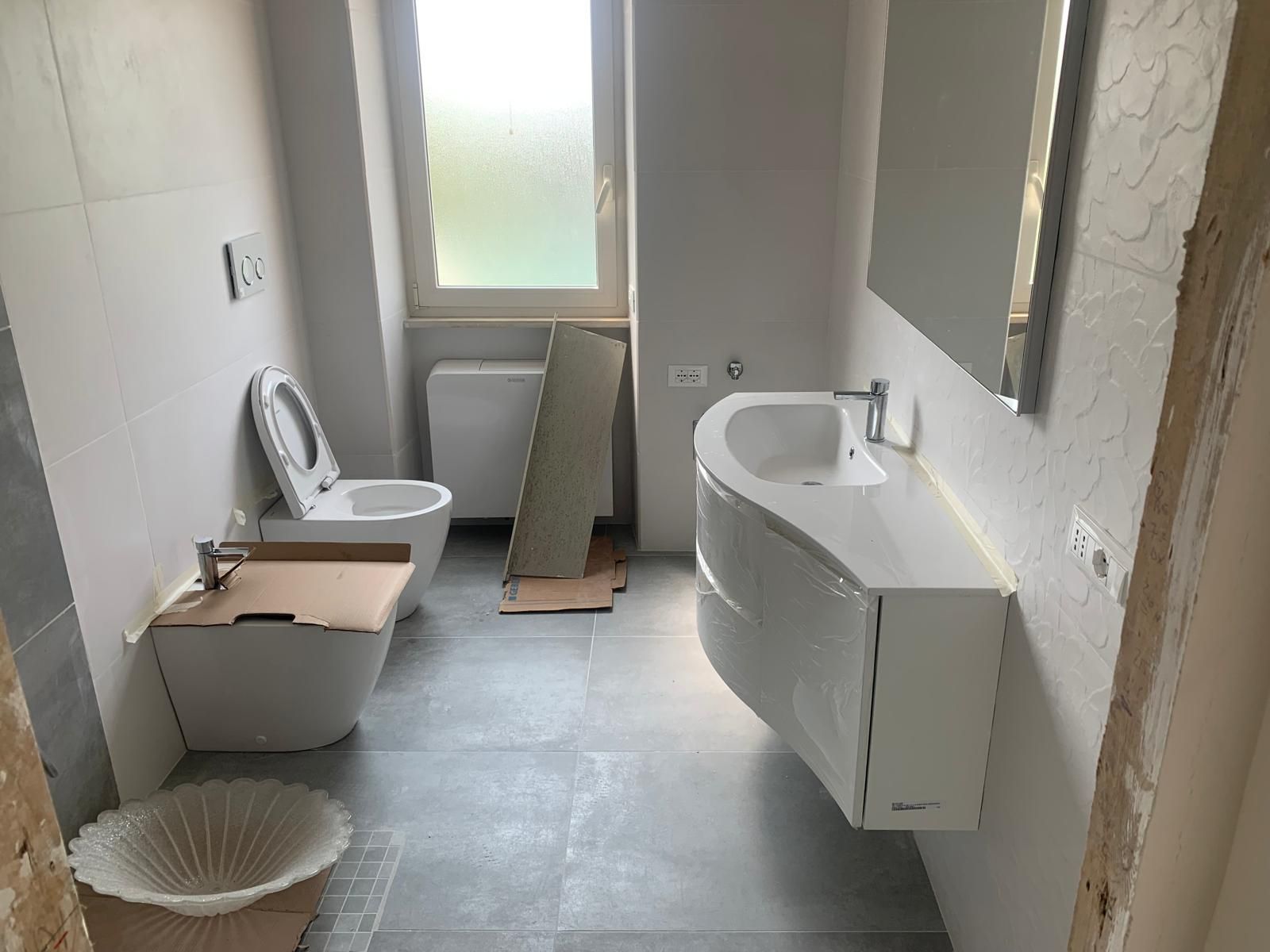 Bagno stretto in fase di ristrutturazione con WC, lavabo, specchio e pavimento piastrellato.