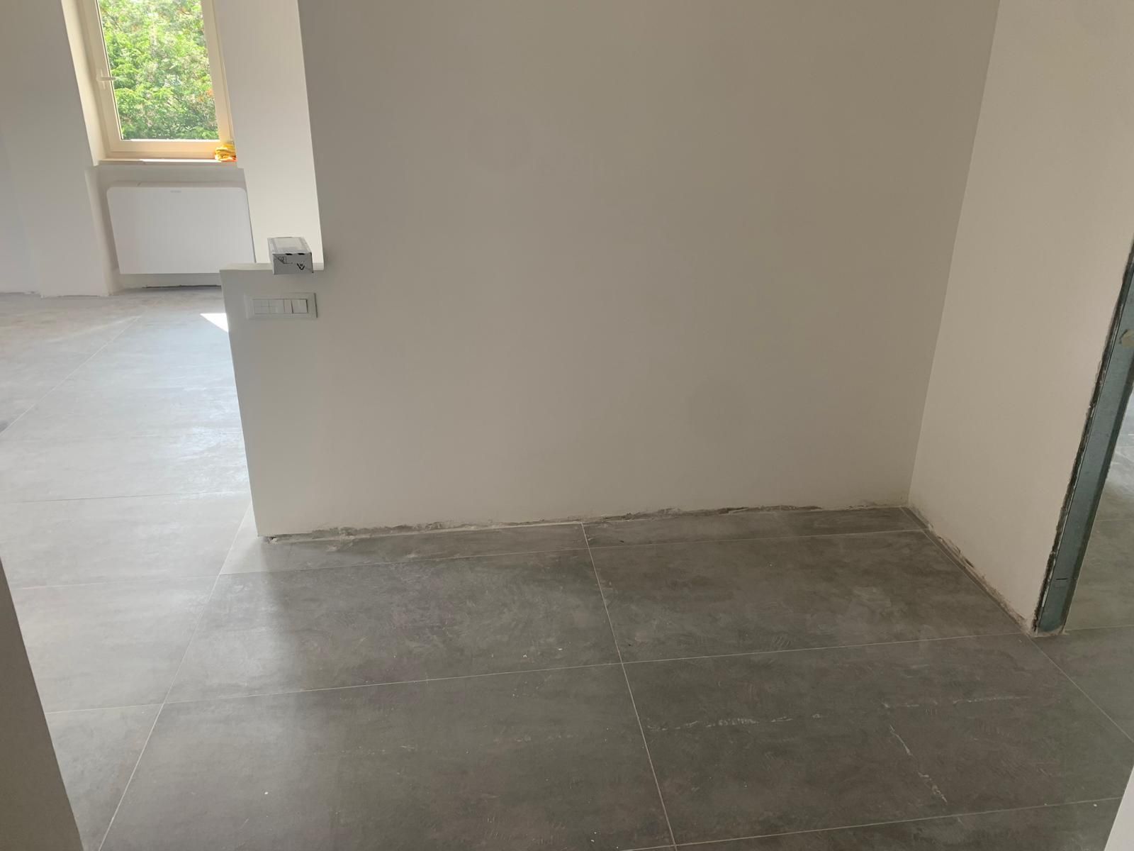 Stanza vuota con pavimento in piastrelle grigie, pareti beige e una porta che si apre su una zona luminosa.