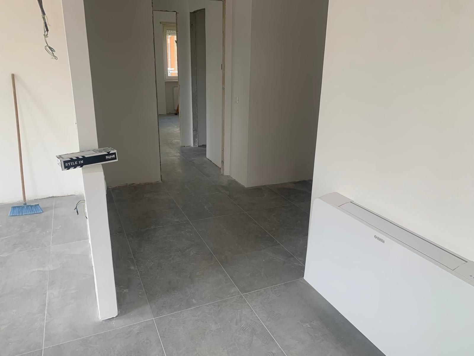 Corridoio vuoto e stanza in ristrutturazione con pavimento piastrellato grigio e pareti bianche.