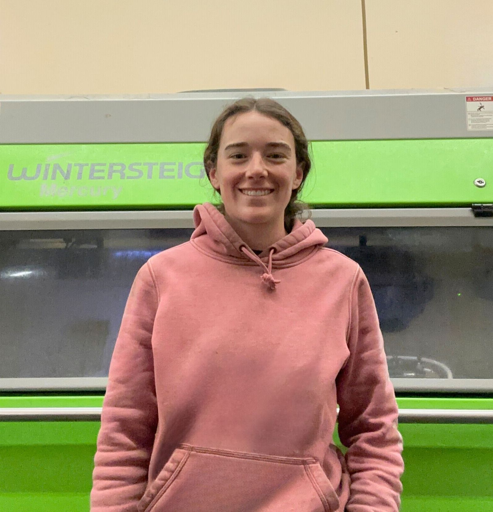 A woman in a pink hoodie is smiling in front of a green machine.