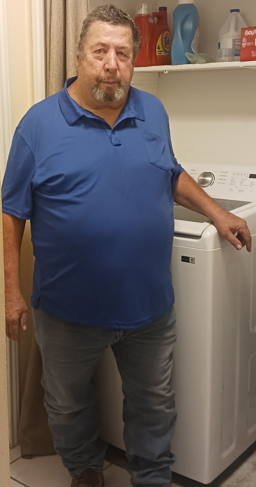 Man in a blue polo stands beside a white washing machine in a utility room.