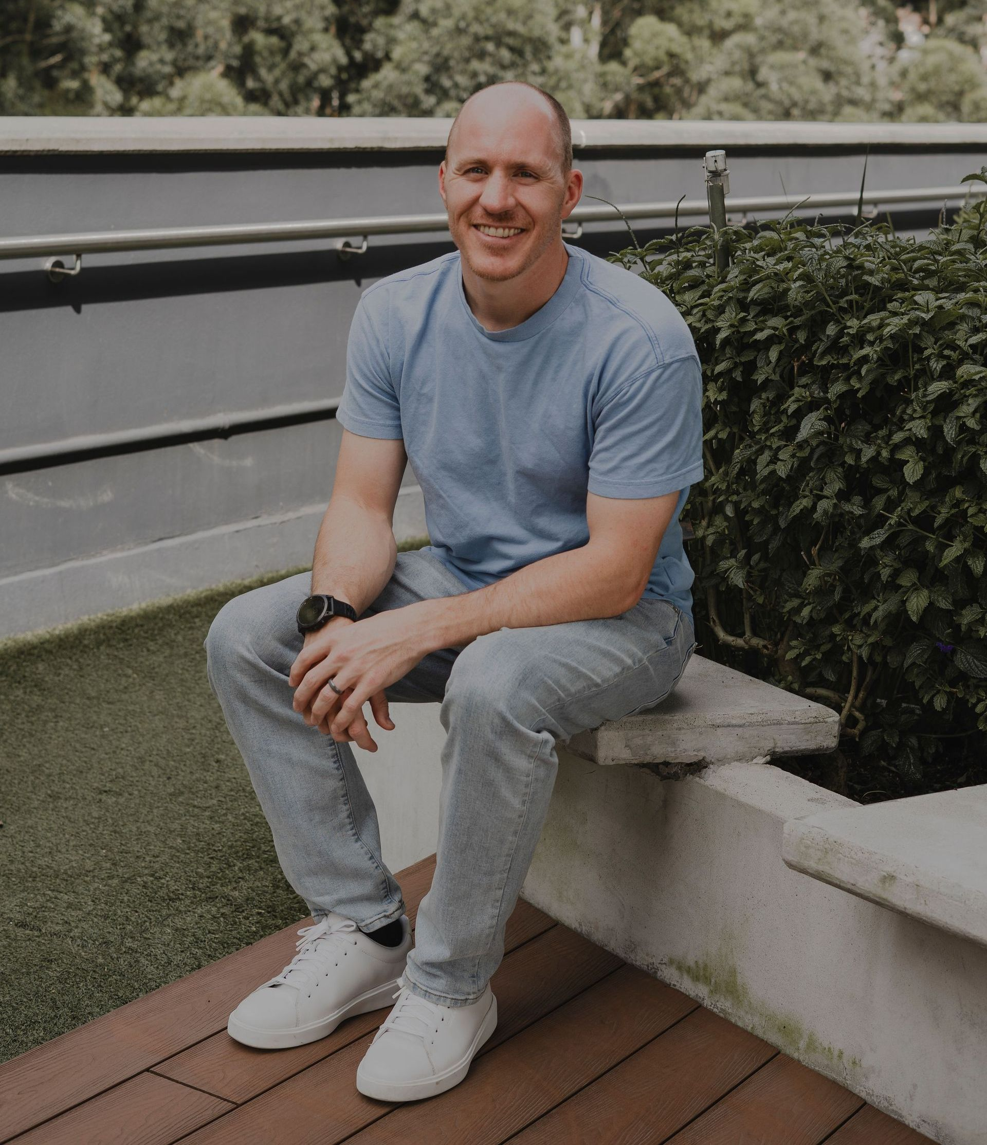 Coach Adam Broda sitting outside, wearing blue shirt, jeans, and white shoes, smiling.