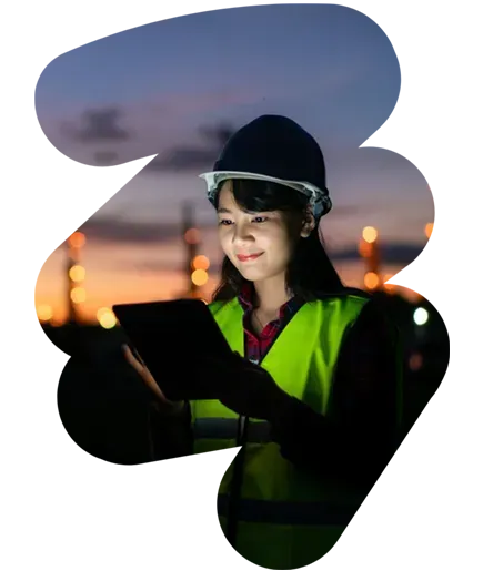 Worker in a hard hat and reflective vest using a tablet at dusk, with blurred city lights behind.