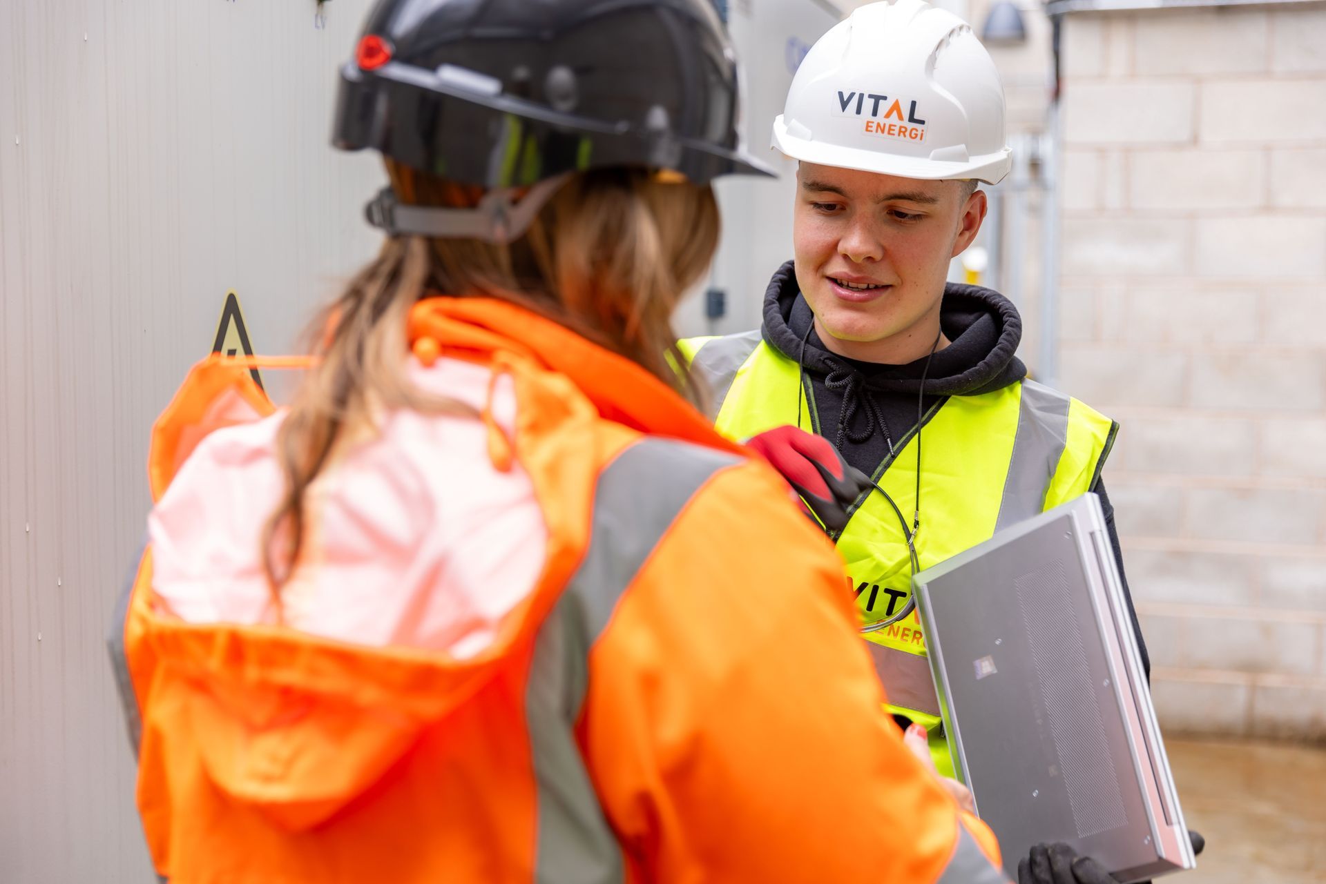 Two construction workers in safety gear discussing plans on a job site, one holding a clipboard.