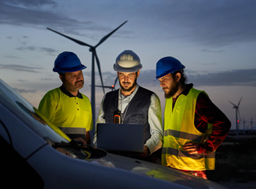 Three engineers in hard hats review a laptop beside wind turbines at dusk.