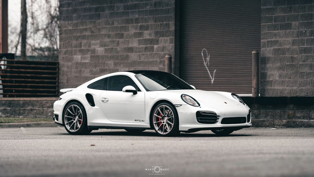 A white porsche 911 turbo s is parked in front of a brick building.