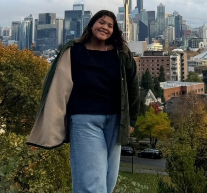 Smiling person standing on a hill with a city skyline in the background, wearing a dark sweater and jeans