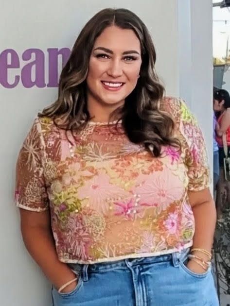 Smiling woman in a pink floral top and jeans, standing indoors with hands in pockets.