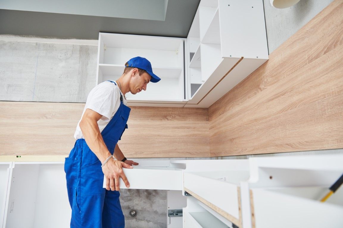 Man in blue overalls installing a kitchen drawer. White and wood cabinets, construction setting.