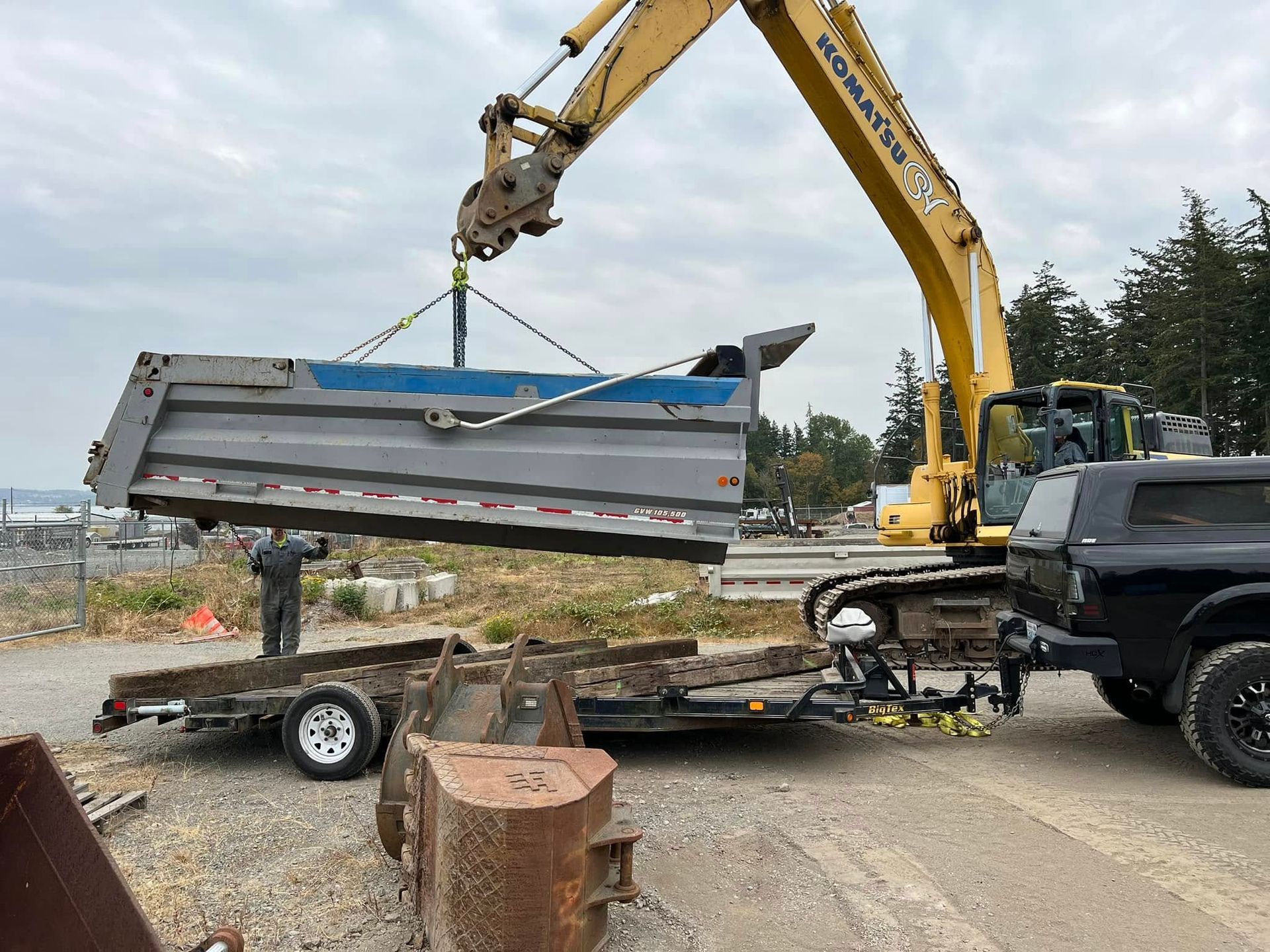 Yellow excavator loading a gray metal trailer bed onto a flatbed truck outdoors